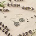Round dog nose print earring laid on textured cream cloth with sprigs of lavender.
