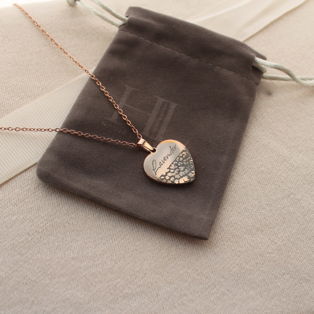 A rose gold heart pendant necklace engraved with Lavender and a textured pattern, resting on a dark gray velvet jewelry pouch over a beige fabric background.
