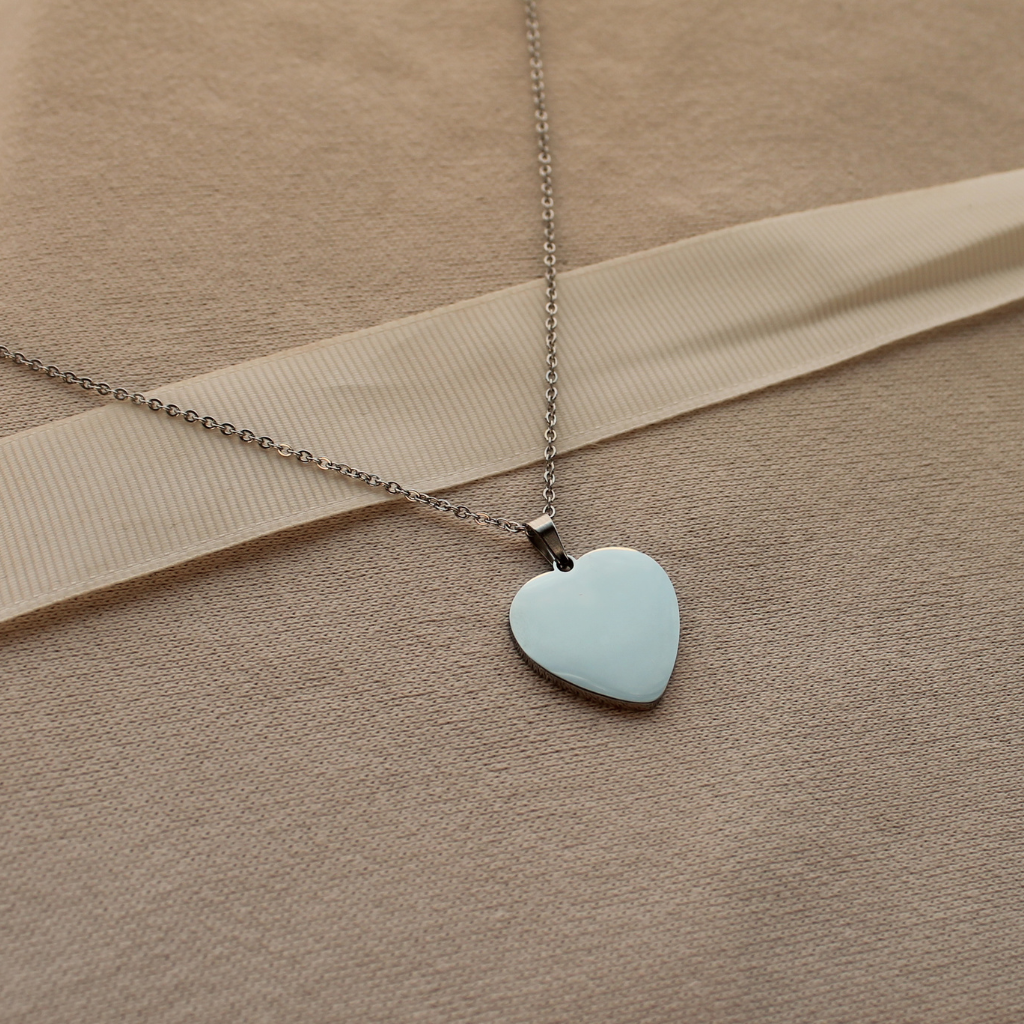A silver heart pendant necklace with a smooth, resting on a beige textured fabric background with a ribbon.