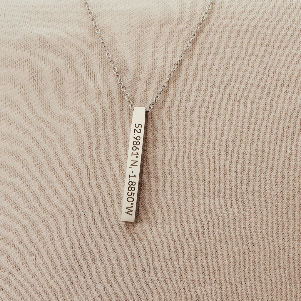 A silver bar necklace with a vertical rectangular pendant engraved with coordinates, displayed on a textured beige fabric background.