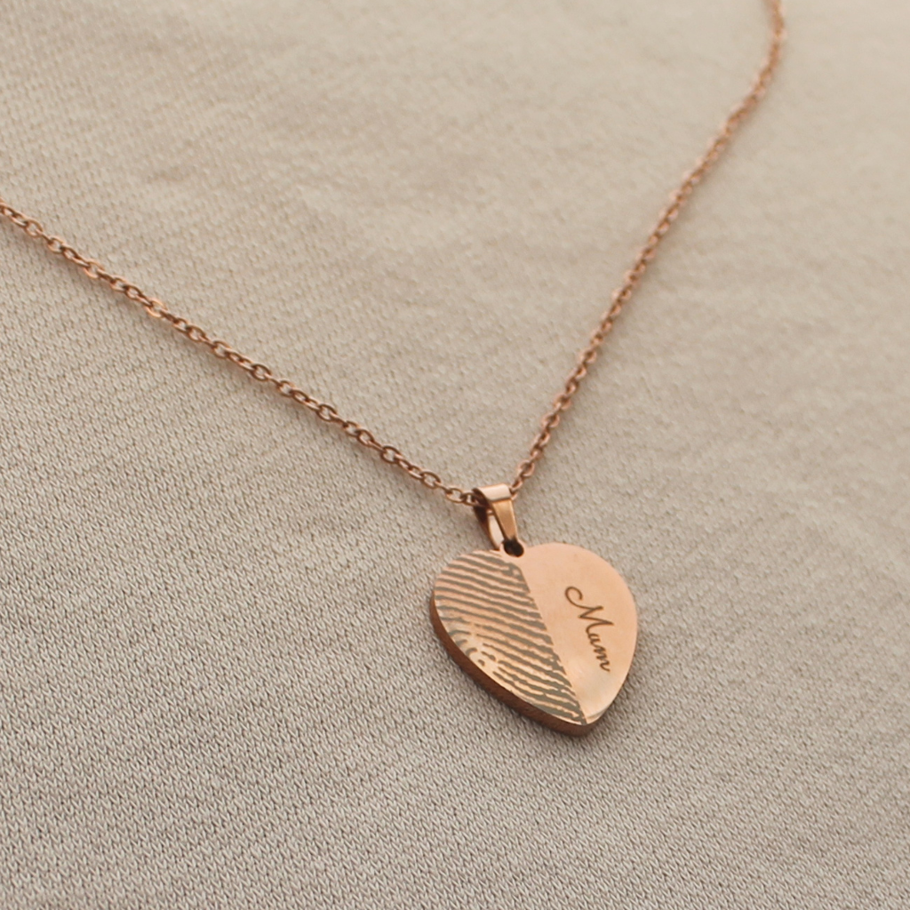 A rose gold heart pendant necklace with a engraved finger print  with 'Mum', displayed on a neutral beige textured fabric background.
