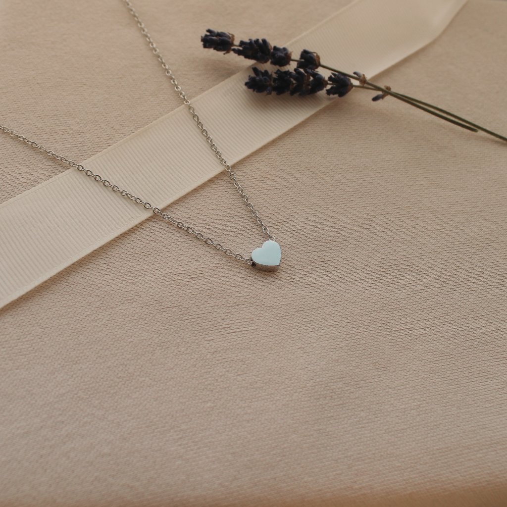 A delicate silver chain necklace with a stainless steel heart-shaped pendant, styled on a beige textured fabric background with a cream ribbon and dried lavender sprigs.