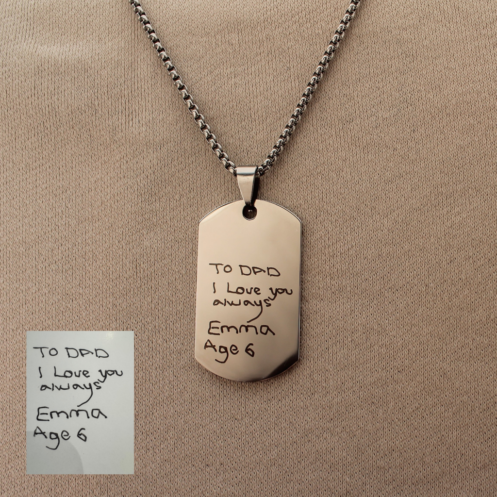 A silver-toned dog tag pendant necklace engraved with 'TO D&D I Love you always Emma Age 6', displayed on a beige fabric background with a small inset photo of the handwritten note.