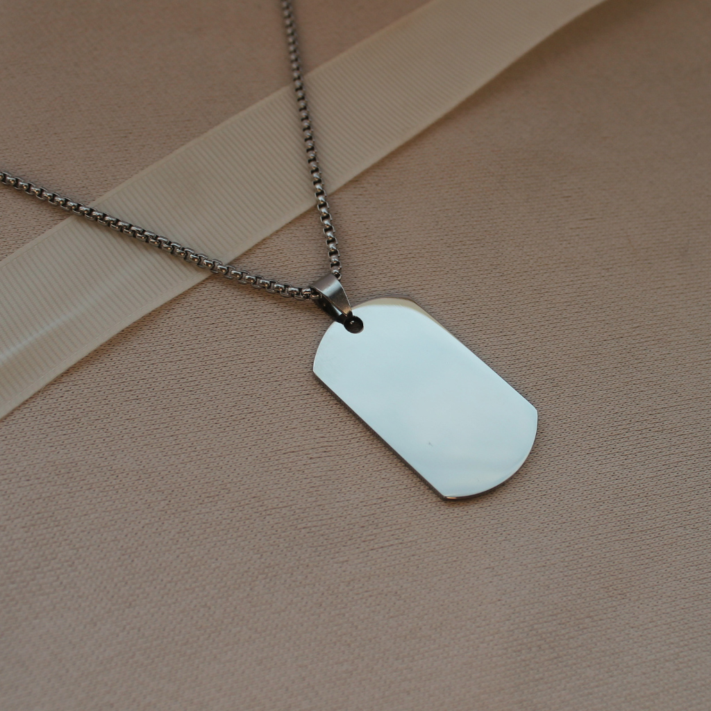 A silver dog tag pendant necklace on a matching silver ball chain, displayed on a textured beige surface with a beige ribbon.