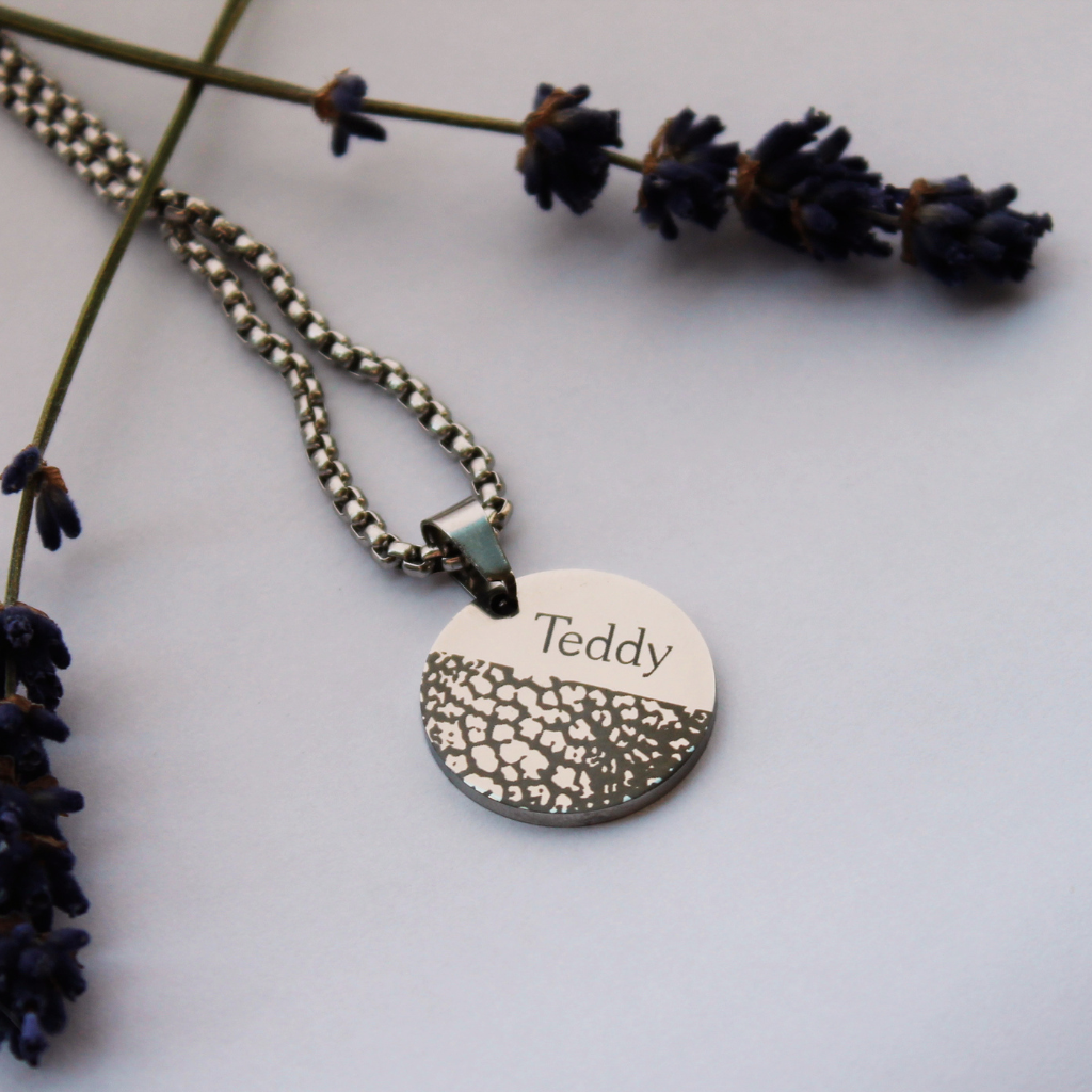 A silver-tone round pendant necklace engraved with the name 'Teddy' and a textured pattern on the lower half, displayed on a light gray background with dried lavender sprigs.