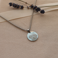 A silver round pendant necklace with a custom engraved dog portrait and the name 'Teddy', displayed on a beige textured fabric with lavender sprigs and a ribbon.