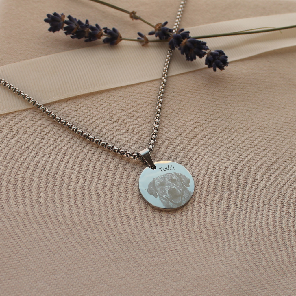 A silver round pendant necklace with a custom engraved dog portrait and the name 'Teddy', displayed on a beige textured fabric with lavender sprigs and a ribbon.
