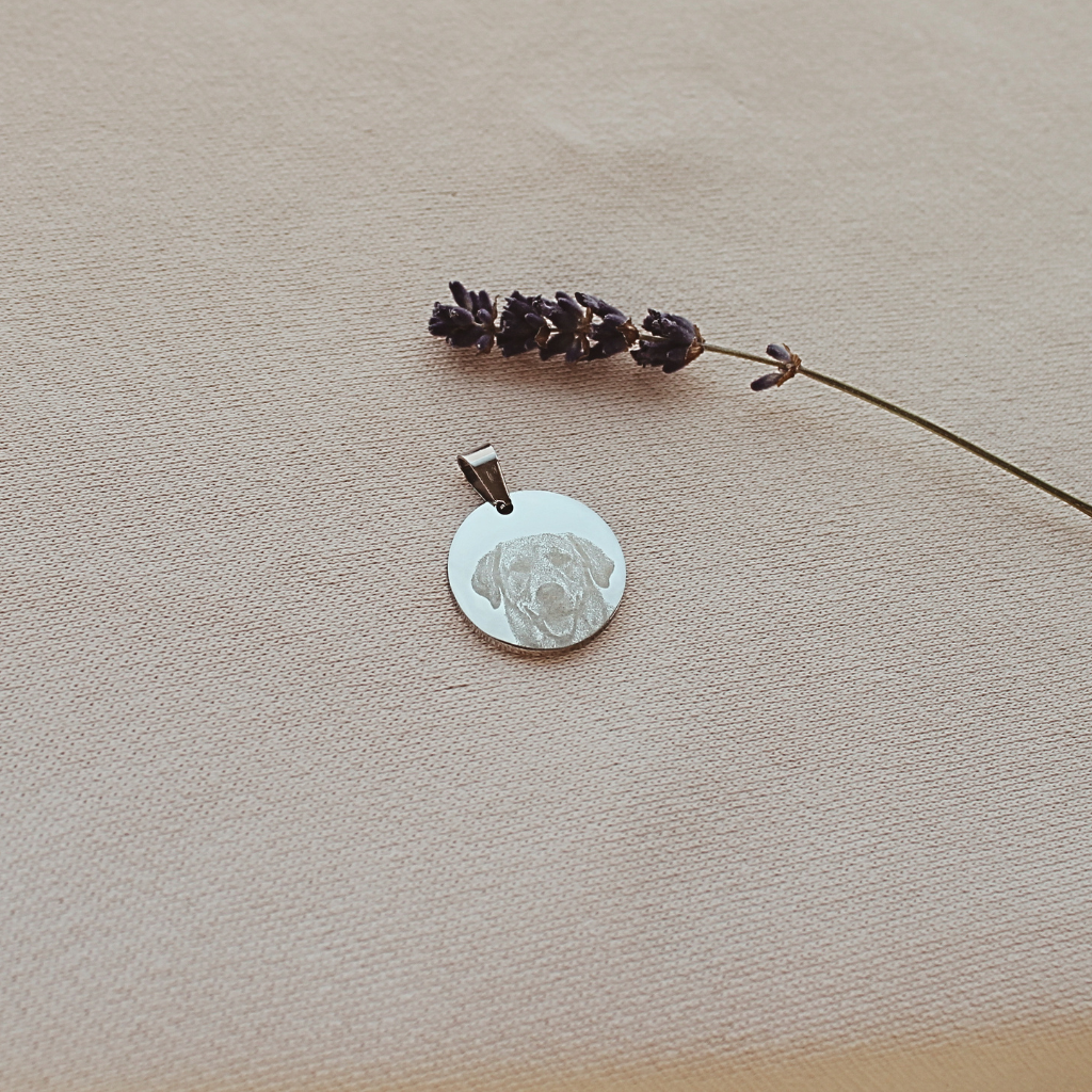 A silver round pendant with an engraved dog face design, displayed on a textured beige fabric next to a dried lavender sprig.