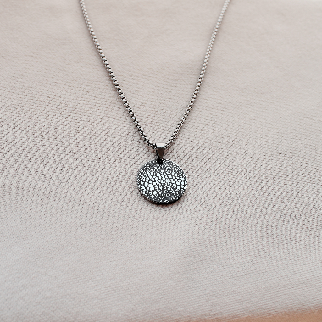 A silver chain necklace with a textured, circular disc pendant, displayed on a soft beige fabric background.
