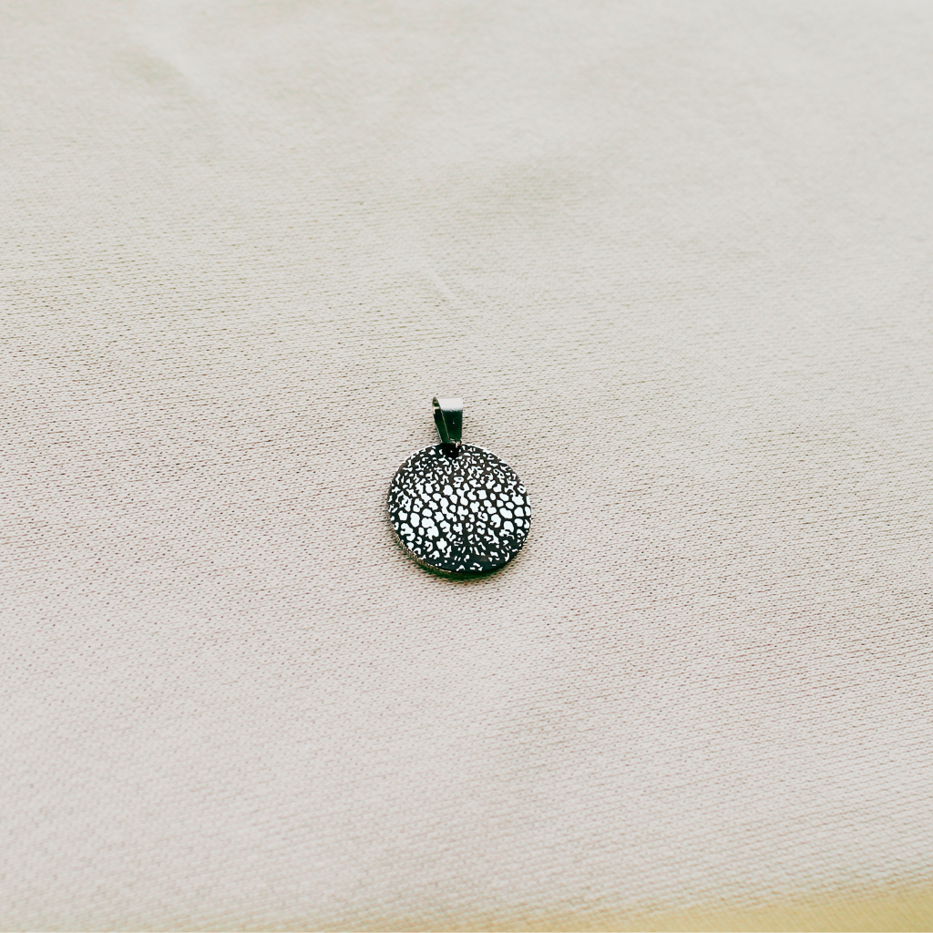 A small round textured pendant in a dark metallic finish, displayed on a light beige fabric background.