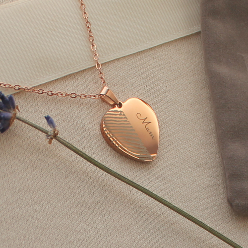 A rose gold heart-shaped pendant necklace engraved finger print and 'Mum'. On a textured beige fabric background, styled with a lavender sprig and a grey pouch.