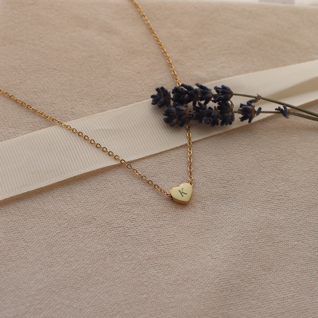 A delicate gold chain necklace with a small heart-shaped pendant engraved with the letter 'K', styled on a beige textured fabric background with a white ribbon and dried lavender sprigs.